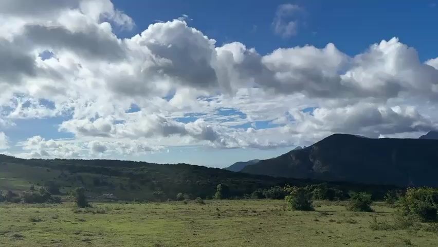 The video shows a landscape with rolling green hills in the foreground and lush green mountains in the background. The sky is blue with patches of white clouds. The clouds are large and fluffy, casting shadows on the landscape. The overall scene is tranquil and scenic. There are no people or animals visible. The camera appears to be stationary.