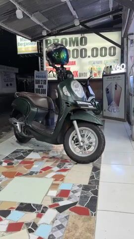 A dark green Honda Scoopy scooter is parked on a colorful tiled floor. The scooter has a brown seat and a black helmet with a rainbow reflective visor is placed on the handlebars. To the left of the scooter, a vertical sign advertises a promotion, stating "PROMO 500.000" and "RAM 12 GB ONLY 900.000". In the background, another scooter is visible and a large sign with text is partially obscured. The lighting appears to be from artificial sources.