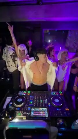 A low-angle shot captures a DJ in a club setting, surrounded by dancing people and bathed in purple light. The DJ, a young man wearing headphones, a cross necklace, and an open white jacket, stands shirtless behind a complex DJ setup with turntables and a digital display. He actively engages with the crowd, smiling and pointing to the music. Several women in white wigs and matching outfits dance behind him, while other partygoers stand around, some in black shirts or patterned clothing. The club's interior is dimly lit, with a few visible details like a decorative wall and an overhead ceiling fixture. The atmosphere is energetic and lively.