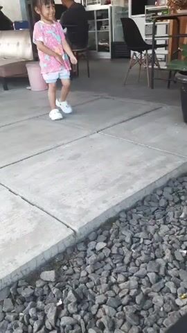 A young girl with black hair and bangs, wearing a pink and blue t-shirt with a star pattern and light blue shorts, stands on a concrete pathway. White Crocs are on her feet. She is smiling and looking to her right, with her left arm bent at the elbow and her hand held near her chest. Her right arm is extended to the side and slightly forward, with her fingers spread. The pathway is bordered by a small area filled with gray gravel. In the background, there is a building with windows, a table, chairs, and a large potted plant. A pink bucket and a stroller are visible on the left side of the image.