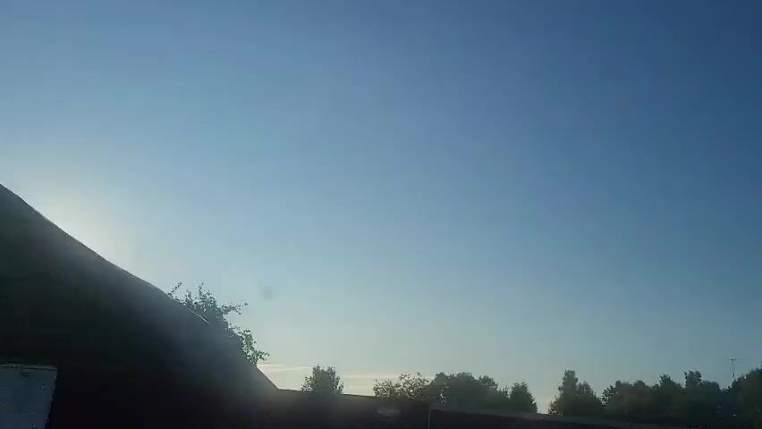 The video shows a bright sun shining from the left side of the frame. The sky is blue and clear with some hazy clouds. In the foreground, there is a dark silhouette of a fence or wall. Behind the fence, there are trees and some vegetation. The video appears to be shot from a moving vehicle, as indicated by the slight camera shake and the changing scenery. There are no people visible in the video. The overall scene suggests a bright, sunny day, possibly in the morning or late afternoon due to the low angle of the sun.