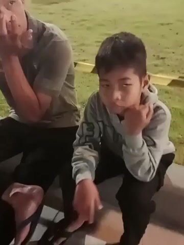 Two males are seated outdoors at dusk. The male in the foreground, a child, is wearing a light gray hooded sweatshirt and dark pants. He is looking down and resting his chin on his hand. To his left and slightly behind him, another male, an adult, is wearing a dark gray t-shirt and dark pants. He is also looking down with his hands clasped near his face. In the background, there is green grass, lighting fixtures, and some yellow caution tape. The child then turns and looks at the adult. The adult then appears to turn away. The child then turns back towards the adult.