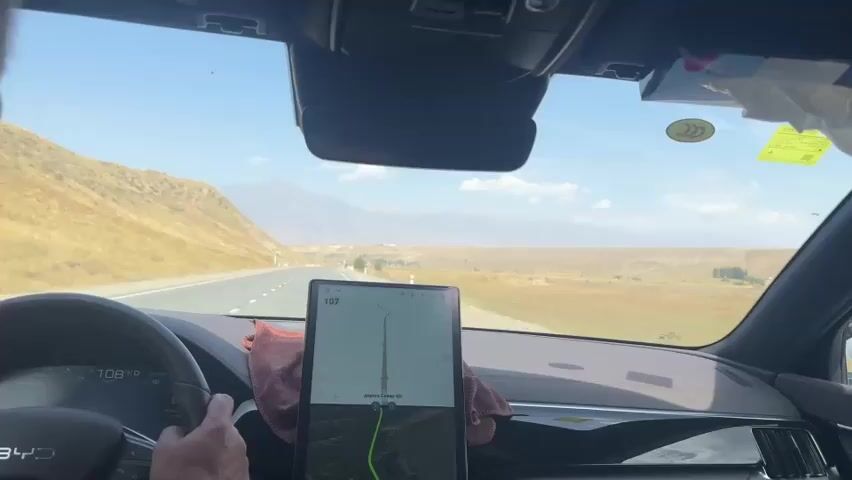 A first-person perspective video captures a journey inside a vehicle, looking out through the windshield and at the dashboard. The driver's hands are visible on the steering wheel, and a large tablet screen displays a GPS navigation system. The exterior view reveals a road stretching into the distance, flanked by dry, rolling hills under a clear blue sky. Mountains are visible on the horizon, and the overall landscape appears arid and sparsely vegetated. The time stamp in the video progresses from 00:00 to 00:04, indicating a brief recording of the drive.
