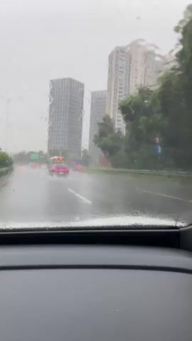 The video captures a view from inside a car driving on a wet road during a rainstorm. Water droplets streak across the windshield, obscuring the view of the cityscape ahead. Several vehicles, including cars, a bus, and a truck, are visible on the multi-lane road, their lights reflecting on the wet asphalt. Tall modern buildings rise in the background, partially obscured by the rain and mist. Green trees line the side of the road. The overall atmosphere is gray and damp, indicative of heavy rain.