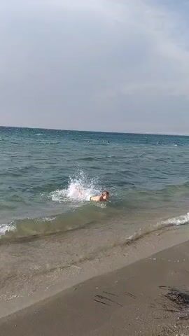 A person is swimming in the ocean near the shore. The water is clear and blue, with waves coming in. The person is wearing white shorts and is swimming on their back. The beach is made of sand. The sky is cloudy.