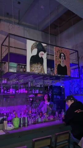 The video shows an interior scene inside what appears to be a bar. The lighting is primarily in shades of purple and blue, creating a mood-setting ambiance. A bartender, a person with dark hair and wearing a denim shirt, is behind the bar, seemingly working on drinks or organizing the area. Two customers are visible: one is seated at the bar wearing a black jacket and a baseball cap, and the other is briefly seen at the edge of the frame.

The bar is well-stocked with a variety of bottles and glasses, neatly arranged on shelves behind the counter. Hanging above the bar, supported by chains, is a metal frame displaying two pieces of art. The artwork consists of two paintings, each depicting a portrait.

The overall setting suggests a relaxed, urban environment, with a focus on art and drinks. The atmosphere feels contemporary and inviting.