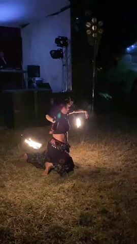 A woman wearing a floral headpiece and a dark, flowing skirt with a patterned belt is kneeling on grassy ground at night. She is holding two flaming torches, one in each hand, and appears to be performing a fire dance. Behind her, there is a stage with lighting equipment, including spotlights and a multi-headed fixture. In the background, other people are visible, and there are structures that suggest an outdoor event or festival. The scene is dimly lit, with the flames of the torches providing a significant light source.
