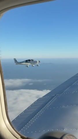 The video shows a view from inside an aircraft window, looking out at another small aircraft flying in formation. The sky is a clear blue, and there are white clouds below. The wing of the aircraft the video is being filmed from is visible in the foreground, with its metallic surface and rivets clearly visible. The other aircraft is white with a single propeller and appears to be a twin-engine plane. The two aircraft are flying at a similar altitude, with the other aircraft slightly to the left and ahead. The scene suggests a formation flying maneuver or a practice flight between two small planes.
