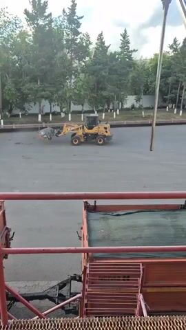 A yellow loader sweeps a paved area. A man is shoveling material into a conveyor belt.