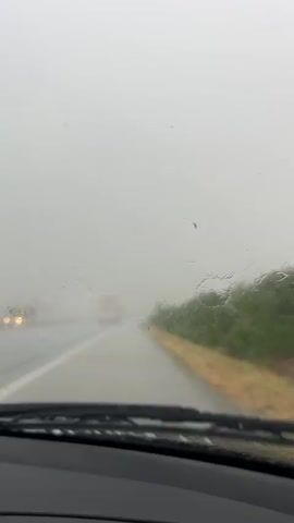 This media file captures a view from inside a car driving on a road during a heavy rainstorm. The windshield is covered in raindrops, distorting the view of the road ahead. The scene is a highway with a lane marker visible on the left and a grassy verge with a dirt shoulder on the right. In the distance, through the fog and rain, faint lights of other vehicles, likely trucks, can be seen. The sky is overcast and a light gray, indicating poor visibility and a gloomy atmosphere. The windshield wipers are active, intermittently clearing a section of the windshield, but the rain is relentless, creating streaks and patterns on the glass. The overall impression is one of a challenging and somewhat isolated drive through adverse weather conditions. The time of day appears to be daytime, but the heavy clouds obscure any direct sunlight. The location context of Zlatina, Bulgaria, suggests a rural or semi-rural highway environment. No text is visible in the media file.
