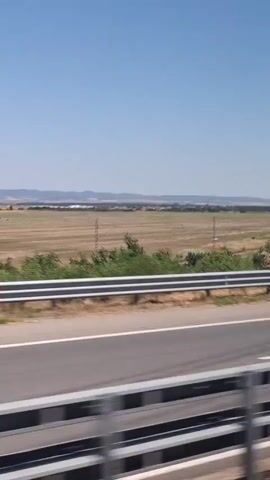 This video captures a view from a vehicle moving along a highway. The perspective is from the passenger side, looking out at a vast, open landscape under a clear blue sky. The immediate foreground is dominated by the highway itself, with multiple lanes visible separated by white lines and bordered by metallic guardrails. Beyond the highway, a wide, flat field stretches towards a distant horizon. The field appears to be a mixture of dry, light brown earth and some patches of vegetation. Several utility poles dot the landscape. On the right side of the frame, a dense cluster of green trees is visible, with some sparse vegetation and bushes closer to the highway. In the distance, a faint outline of hills or mountains can be seen. The overall impression is one of a sunny day in a rural or semi-rural environment. There are no people clearly visible in the frame.