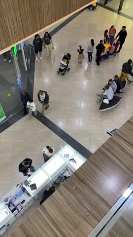 The video shows a wide shot from above of a shopping mall atrium. Several people are milling about, some walking, others sitting. In the foreground, there is a retail counter with what appears to be electronics for sale, such as phones and computers. A person in a black jacket is standing behind the counter. Further back in the atrium, several individuals are gathered around a stroller, and a small group is sitting on a curved bench. A child is riding in a toy car with a handle, being pushed by an adult. The mall has polished tile floors and wooden accents. There are large glass windows, suggesting an indoor environment. The lighting is bright, indicating it is daytime. No specific time of day or weather conditions can be determined. There are no visible signs with text. The overall atmosphere appears to be one of casual shopping and everyday activity.