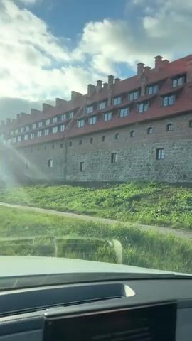 The video shows a long, multi-story building with a red tiled roof and brick and stone walls. The building appears to be a castle or a large historical structure. There are many windows with dark frames visible on the upper floors. The building is situated on a grassy hill, and a path is visible leading up to it. The sky is blue with scattered white clouds, and the sun is shining, casting a light on the building. The footage appears to be taken from inside a vehicle, as indicated by the presence of a car dashboard in the foreground. There are no people visible in the video.