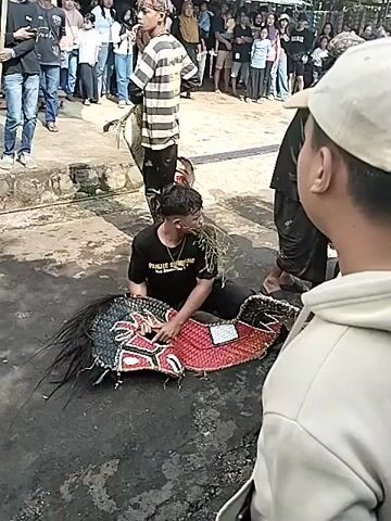 The video shows a man in a black t-shirt sitting on the ground, holding and chewing on some straw. He is next to a large, colorful woven mask with a horsehair mane. The mask is red, black, and white. There are other people standing around, watching. The ground appears to be wet and there is a concrete surface in the background. A crowd of onlookers is visible further back. One man is walking past the group.