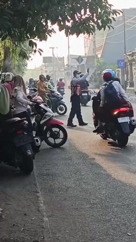 A street scene shows several motorcycles parked on the side of the road. A traffic officer in an orange vest and a blue cap is directing traffic. He is holding his right arm out and waving his hand. Several people are on motorcycles, some of whom are wearing helmets and some are wearing headscarves. The officer is facing to the right of the frame, and several motorcycles are moving from left to right in front of him. A stop sign is visible in the background on a pole. The sun is shining, casting long shadows.