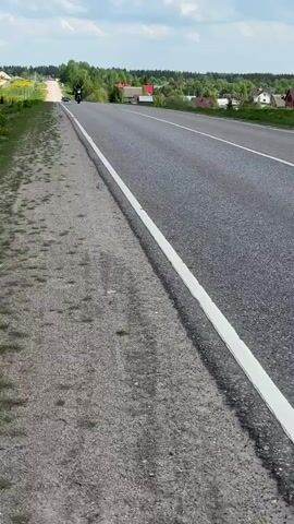 The video shows a person riding a motorcycle on a paved road. The road is straight and has a white line marking the edge of the lane. The road has an unpaved shoulder. The rider is wearing a black helmet and dark clothing. In the beginning of the video, houses are seen in the distance beyond the road. The sky is blue with fluffy white clouds. There is a car in the distance. Later in the video there is a field and then trees. The motorcycle passes the camera and then disappears into the distance.
