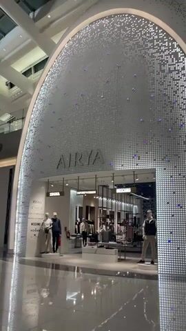 The video begins with a medium shot of a store entrance labeled "AIRYA". The store's facade is decorated with a white, mosaic-like pattern interspersed with small blue lights. Inside the store, mannequins display clothing, and racks of garments are visible. To the left of the store, another mannequin is dressed in an off-white outfit. To the right of the store, a mannequin stands wearing a dark vest over a light-colored shirt and dark pants. The camera then pans to the right, showing a pillar with an illuminated white patterned surface. Further to the right, a sign with yellow and green elements is visible, along with a poster for "CAFE". The video then pans left, revealing a wide shot of an airport terminal interior. People are seen walking, some with luggage. A woman in a red top and a person in glasses with a pink suitcase are seated. Other individuals are also seated in the waiting area, with luggage around them. The environment appears to be modern and spacious, with high ceilings and polished floors reflecting the overhead lighting.