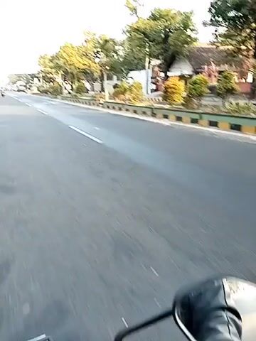 A video captured from a moving vehicle shows a road with a center line. On the right side of the road, there are trees, plants, and a building. A truck is visible with its trailer tilted at an angle, as if it has tipped over or is in the process of doing so. It appears to be partially entangled with the trees. On the left side of the road, there is traffic, including cars and motorcycles. The sky is clear and bright, suggesting it is daytime.
