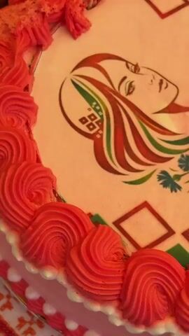 The video shows a close-up of a cake. The cake is decorated with an edible image featuring a stylized drawing of a woman with long, flowing hair in shades of red, green, and white. Her hair is adorned with a pattern that resembles traditional Belarusian embroidery. A sprig of blue flowers with green leaves is placed near her neck. To the right of the woman's image, text is visible in Russian: "ГОД БЕЛОРУССКОЙ ЖЕНЩИНЫ 2026," which translates to "YEAR OF THE BELARUSIAN WOMAN 2026." The cake itself is pink, and its border is decorated with swirls of red frosting. The background is indistinct.