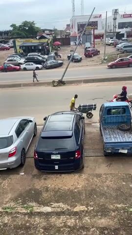 This media file captures a bustling street scene in Ogbunike, Nigeria, under an overcast sky. The foreground features several parked cars, including a silver sedan and a dark blue SUV. Beyond these, a busy road stretches into the distance, lined with buildings and more parked vehicles. A trio of people are visible on the side of the road, one on a motorcycle, and two individuals with a cart. Further down the road, several cars are in motion, and a yellow motorized tricycle, a common mode of transport in the region, is navigating the traffic. The overall impression is one of daily urban life with economic activity. The time of day appears to be daytime, with the cloudy weather suggesting it might be a humid or rainy season. Visible text includes "THE NEW 440ML" on a large billboard, and what appears to be "UBA" on a building facade, possibly indicating a bank. The scene conveys a sense of vibrant, everyday activity.
