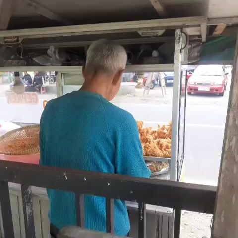 A person with gray hair, wearing a blue sweater, stands with their back to the camera at a food stall. The stall has a glass display case filled with fried food. To the left, a mesh basket containing more food is visible. Behind the stall, a street with vehicles and buildings can be seen. The person appears to be tending to the food.
