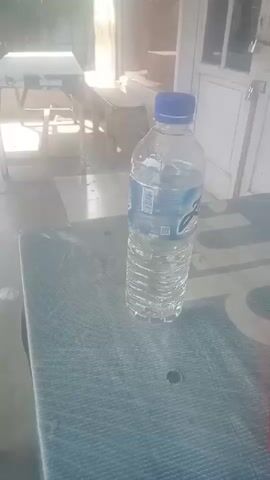A clear plastic bottle filled with water, with a blue cap, stands upright on a blue patterned table. To the right of the bottle, a red cylindrical object, possibly a pen or marker, lies on the table. The background is blurred, showing what appears to be furniture and a door. The scene is outdoors, with daylight filtering in.