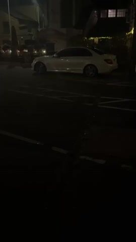 A white sedan is parked on the side of a dark street. The car is angled slightly towards the viewer. The street is paved and appears to be a road with parking spaces marked by white lines. There are buildings in the background, and some lights are visible, suggesting it is nighttime or early morning. The overall lighting is dim. The video is approximately four seconds long and shows no people or discernible actions.