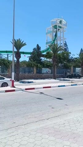 The video shows a water slide structure with a tall white tower and a green slide against a clear blue sky. Palm trees and other greenery are visible in front of the structure, behind a fence. A sidewalk with red and blue striped borders runs along the foreground, with an asphalt road beyond it. A white car is parked on the left side of the road, and a black car is parked on the right. A light pole stands on the left side of the frame. The scene appears to be a sunny day at a water park or resort.