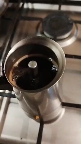 The video shows a close-up, overhead shot of a moka pot on a gas stovetop. The moka pot is made of stainless steel and has a shiny exterior. Coffee is actively brewing in the top chamber of the moka pot, with bubbles forming on the surface and the dark liquid rising up the central spout. The stovetop is also made of stainless steel and has a black circular burner visible in the background. The lighting is dim, suggesting it may be evening or indoors. The video is very short, lasting only a few seconds, and focuses entirely on the coffee brewing process.