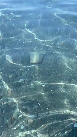 The video shows a top-down view of shallow, clear blue water. The water is calm with gentle ripples and reflections of light playing on the surface. Beneath the water, a dark, pebbly seabed is visible. The pebbles are of various sizes, mostly dark gray and black, with some lighter-colored ones interspersed. The light from above creates dappled patterns and bright streaks on the seabed. The video appears to be recorded at a low angle, close to the water's surface, giving the impression of looking down from a boat or the edge of a shore. There are no people or prominent objects visible in the frame, only the natural environment of the water and seabed.
