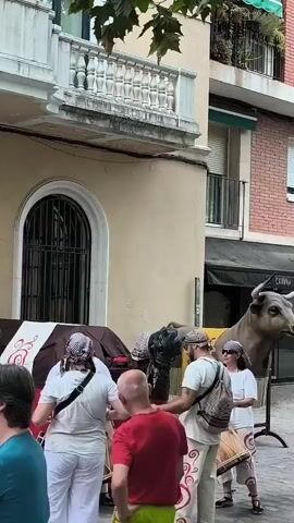 The video shows a group of people gathered on a street in front of buildings. A large, faux cow sculpture is present to the right of the frame. Several people are wearing white clothing and some have bandanas on their heads. Two individuals are holding and appears to be playing drums. One person is wearing a red shirt and bright green shorts. The background consists of buildings with balconies and windows, suggesting an urban setting. The lighting indicates it is daytime.