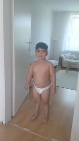 A young boy stands in a doorway, wearing only a diaper. He is smiling and appears to be happy and energetic. The boy is the main subject of the video, and his movements and expressions are the focus. The background shows an indoor setting, likely a home, with a hallway and a living room visible. The lighting suggests it is daytime. The scene appears to be casual and domestic, with no indication of a formal event or performance. The boy's actions suggest he might be playing or dancing.
