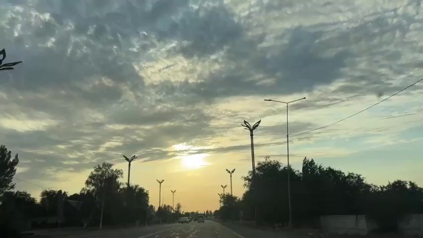 The video shows a view from inside a vehicle traveling on a road at sunrise. The sky is filled with dramatic clouds, and the sun is visible low on the horizon, casting a warm glow. Several streetlights with distinctive decorative tops line the road, along with trees and some buildings. Other vehicles are also seen on the road, suggesting a somewhat populated area. The overall impression is of a calm and scenic morning commute.