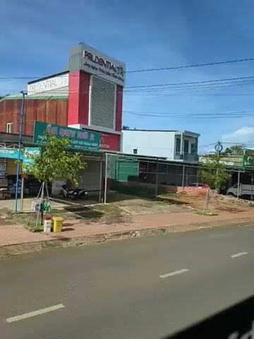 The video shows a street scene, likely in a developing country, on a bright, sunny day with a clear blue sky and some scattered clouds. The camera is moving, suggesting it is attached to a vehicle. On the left side of the road, there are several buildings with storefronts. The first building visible has a red and white sign that says "PRUDENTIAL" in large letters, along with Vietnamese text. Below this, there's another sign with "PRUDENTIAL" and Vietnamese text. To the left of the Prudential building, there's a smaller storefront with a green awning and a motorcycle parked in front.

Further down the street, other businesses are visible, some with green awnings and Vietnamese signage. One building has a sign that appears to be for a taxi service. Another sign reads "MAN PHUONG THAO." There are various vehicles, including a white truck, a small silver car, and a yellow bus with cartoon characters (Pikachu) on its side, indicating it's likely a passenger bus. The bus has "LUXURY 34 PHONG" written on it. Some buildings are open-air structures, and there are various objects like carts, trailers, and plants visible along the sidewalk. Power lines are strung across the top of the frame. The road itself is paved, with white lane markings. The overall impression is of a commercial area with small businesses and traffic.
