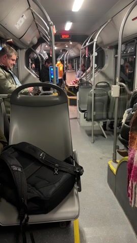 The interior of a public transit bus is shown. The bus is moving, with visible motion and sound. Passengers are standing and sitting. A black backpack rests on the seat in the foreground. The walls and ceiling are light gray. Handrails are visible throughout the bus. A red "STOP" sign is illuminated at the front. Several people are standing near the front of the bus. A machine for purchasing tickets is visible.  A multicolored scarf is draped over a seat.
