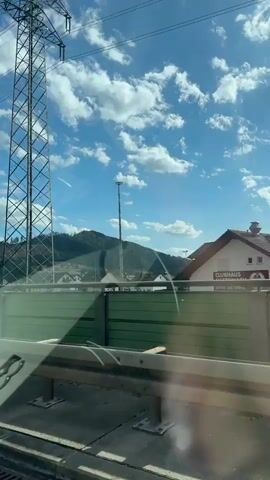 A view from a vehicle traveling on a road. In the foreground, a green sound barrier runs along the side of the road. A metal guardrail is visible in front of the barrier. In the middle ground, a transmission tower stands tall, and a smaller tower with a light is also visible. Beyond the towers, a tree-covered hill is visible with some areas of deforestation. The sky is blue with scattered white clouds. A reflection of the car's interior, including a side mirror and part of the dashboard, is visible on the windshield. The reflection shows what appears to be a Fiat 500 logo.