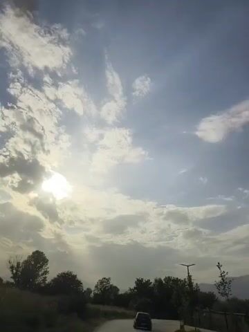 The video shows a view from inside a moving vehicle. The sun is partially obscured by clouds, casting rays of light through the sky. The road curves to the right, with trees and vegetation lining the side. A dark-colored car is visible in front, also on the road. The overall scene suggests a daytime drive in a rural or suburban area with dramatic cloud formations.