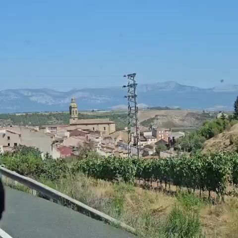 The video shows a panning shot of a village with a prominent church. The church has a bell tower with multiple levels and a spire. The buildings in the village are mostly made of stone and tile. In the background, there are rolling hills and mountains. The sky is clear and blue. The overall impression is of a rural, sunny day.