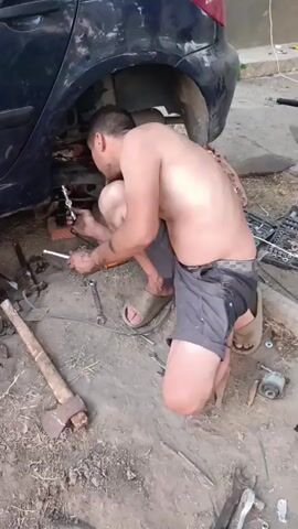 A shirtless man wearing shorts and sandals is kneeling on the ground, working on the underside of a dark-colored car. He is holding a wrench and appears to be tightening or loosening a bolt. Several tools are scattered around him on the dirt ground, including an axe, various wrenches, and what appears to be part of a car suspension. The car is parked outdoors, with a building visible in the background. The man’s posture suggests he is in a somewhat awkward position to access the car's undercarriage.