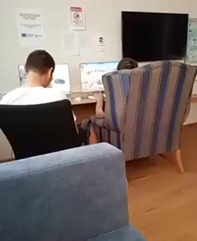 Two individuals are seated at separate computers in a room with light-colored walls. The person on the left is facing away from the camera, looking at a laptop with a white screen displaying what appears to be a video or image. They are wearing a white t-shirt and dark pants. The person on the right is seated in a striped armchair, also facing away from the camera, and is looking at a computer monitor. Their legs are visible, and they appear to be wearing dark shorts. The monitor in front of them shows what looks like a video game with landscape imagery. In the background, there is a large black television mounted on the wall, a wooden table, and several posters or documents attached to the wall. A blue sofa is visible in the foreground, partially obscuring the view. The flooring is made of wood. The overall lighting suggests an indoor environment.