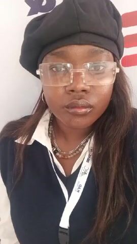 A person with dark skin, wearing a black beret, clear-framed glasses, a white collared shirt with a navy blue sweater vest, and two necklaces is looking at the camera. The person moves their right hand to their ear. They have long dark hair. They are wearing an event lanyard. There is a white wall with a red and blue stripe in the background.