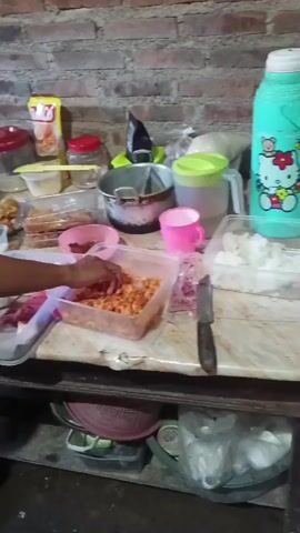 The video shows a dimly lit kitchen with brick walls. In the foreground, a person's hand is seen reaching for a container of food on a table laden with various containers of ingredients, kitchen utensils, and food items. The person is wearing a green and white patterned garment and a dark headscarf. Another person, also wearing a headscarf and a green and floral patterned garment, is visible in the background. Various cooking pots and containers are placed on shelves and counters, suggesting food preparation is in progress. The floor appears to be dirt or concrete.