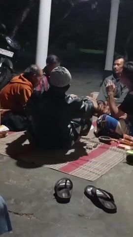 A group of men are gathered outdoors at night, sitting on mats on the ground around a small collection of items including cigarettes, possibly food packaging, and a small cup. The men appear to be in conversation, with some of them smoking. One man in the foreground is wearing a black jacket and a white cap, facing away from the camera. Another man to his right is wearing a gray hoodie and is looking towards the man in the black jacket, holding a cigarette to his mouth. To the left of the man in the black jacket, a man in an orange hoodie is also seated. The environment suggests a casual outdoor setting, possibly a porch or patio, with pillars visible in the background and dark foliage beyond. Two pairs of flip-flops are visible on the ground in front of the seated group. The overall impression is of a relaxed social gathering.