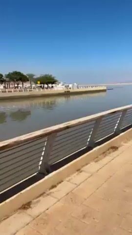 The video shows a waterfront scene on a bright, sunny day. A paved walkway with a low railing runs alongside a body of water, likely a bay or harbor. The water is calm and reflects the clear blue sky. Across the water, a shoreline is visible in the distance with some structures and vegetation. A few people can be seen walking along the waterfront promenade.