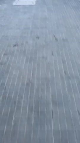 The video shows an urban scene in Sant Vicenç dels Horts, Spain, during daylight hours.

It begins with a blurry shot of a street with a yellow line and a gridded metal surface, suggesting rapid movement. A person in a black jacket and green pants walks past, carrying a red and brown bag. The camera then focuses on this bag, which has "CARBÓN CARVAG" printed on it with a flame design, being held by a hand. The scene shifts to a blurry view of buildings and a street, then focuses on a man with a beard, wearing a black vest, white t-shirt, and blue pants, walking on a sidewalk. He is carrying a yellow plastic bag and smiles at the camera. A red brick building is visible in the background, and another person is partially seen walking alongside him.