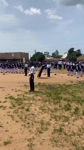 The video shows a group of people in uniform standing in formation outdoors. The person in the foreground is wearing a white shirt and dark pants and is holding a Nigerian flag. Behind them, a larger group of people in similar uniforms are lined up. The environment appears to be an open, dusty area with some grass. Buildings and trees are visible in the background. The sky is cloudy. The overall scene suggests some kind of ceremony or parade.