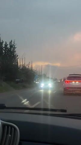 The video captures a first-person view from inside a car, looking out through the windshield onto a road. The time of day appears to be either dawn or dusk, as the sky features soft yellow, orange, and pink hues under an overcast atmosphere. The setting is a semi-rural area, possibly Sidi Abdallah Ghiat, Morocco, characterized by bare trees, some foliage, and power lines running alongside the road.

As the car drives forward, a large, colorful, cartoon-like head sculpture, predominantly blue and pink, is briefly visible on the left side of the road. Other vehicles are present, including a white SUV with illuminated red taillights driving ahead in the same direction, and a car with bright headlights approaching from the opposite lane. The car's dashboard and a portion of the rearview mirror are visible in the foreground. No specific text is clearly legible, though a car's license plate in the distance is too blurry to read. The overall scene suggests movement along a somewhat quiet highway at twilight.