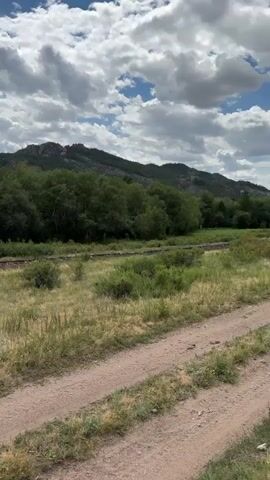 A dirt road cuts through a grassy field. Beyond the field is a dense line of trees, and behind the trees is a mountain range. The sky is filled with white, puffy clouds. The camera pans slowly from left to right, revealing more of the mountain range and the sky.