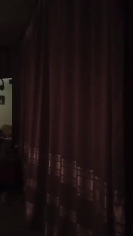 The video captures a dimly lit room, likely a bedroom or living space, in Semey, Kazakhstan. The scene is characterized by its low light conditions, suggesting it is either early morning, late evening, or the curtains are drawn against daylight.

The primary subjects are indistinct objects within the room. A bed is visible, partially covered with bedding, and appears to be the central focus of one area of the room. Nearby, there is a piece of furniture, possibly a dresser or a desk, with items placed upon it, though their specifics are obscured by the low light. In another part of the room, a curtained window allows a strong beam of light to enter, illuminating a section of the space and highlighting the texture of the curtains, which are a muted reddish-pink color. There is also what appears to be a framed picture hanging on the wall. In the background, a doorway or archway leads to another, darker area of the dwelling.

The video appears to be a slow, panning shot, moving across the room. There is no discernible activity or event taking place, beyond the act of recording the space. The mood is quiet and somewhat subdued, due to the low lighting. The location cues, such as the mention of Semey, Kazakhstan, place the scene geographically. No text is visible in the video.