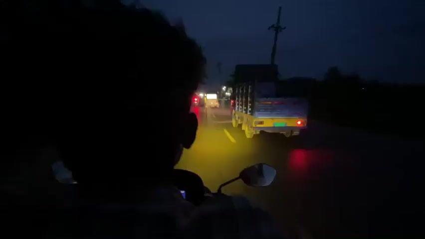 A person is riding a motorbike in the dark. The motorbike's headlight illuminates the wet road ahead. Other vehicles, including a truck, are visible in the distance, with their headlights and taillights glowing. Buildings and trees line the sides of the road.